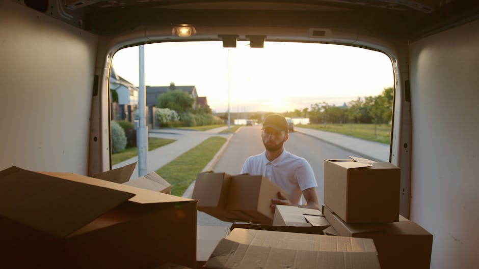 A man with a beard, wearing a blue beanie, a navy and red waterproof jacket, black pants, and white sneakers, is seated on the edge of an open van trunk. Inside the van, several large cardboard boxes 
